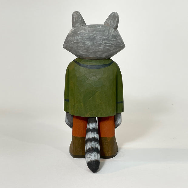 Back angle of small painted wooden sculpture of a sweet looking raccoon, standing and wearing a green coat, yellow and brown shirt, burnt orange pants and brown boots. Its striped tail can be seen.
