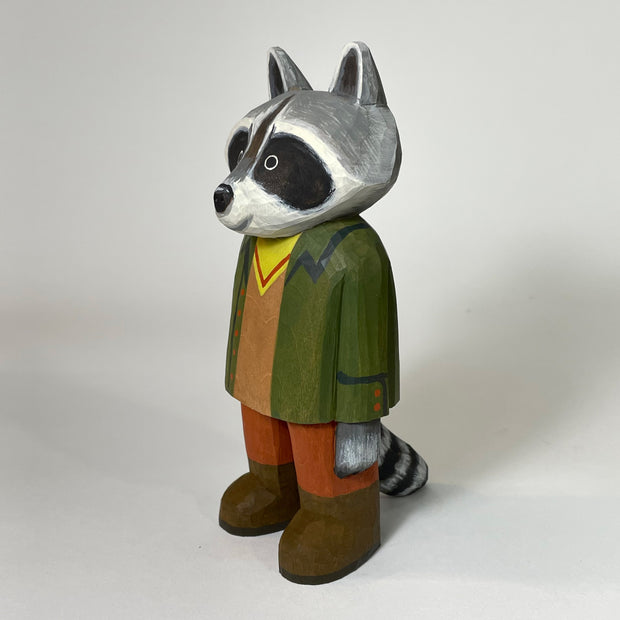 Side angle of small painted wooden sculpture of a sweet looking raccoon, standing and wearing a green coat, yellow and brown shirt, burnt orange pants and brown boots. Its striped tail can be seen.