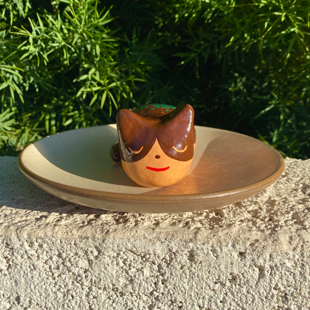 Sculpture of a sleeping tan cat, with simplistic facial features and a red smile. Cat has darker brown accent coloring towards the top of its body, with green flecks on its back to make it resemble a takoyaki.