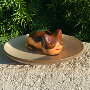 Sculpture of a sleeping tan cat, with simplistic facial features and a red smile. Cat has darker brown accent coloring towards the top of its body, with green flecks on its back to make it resemble a takoyaki.