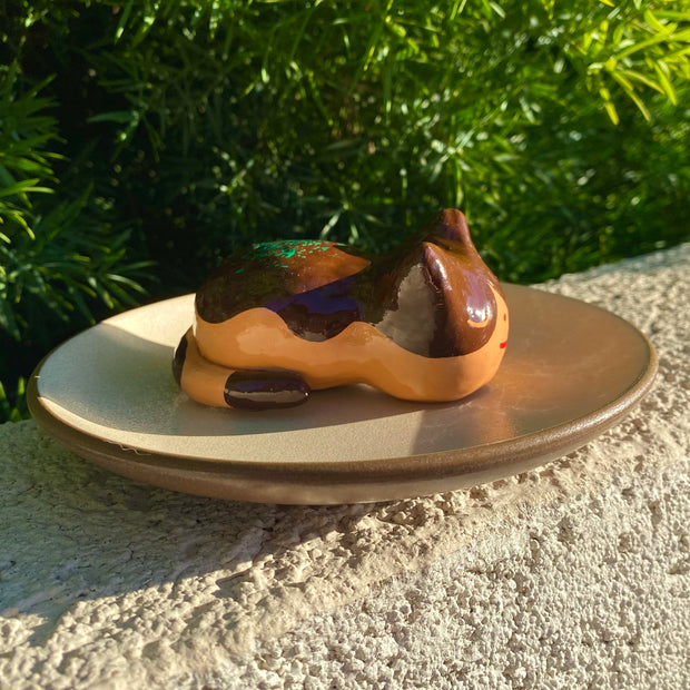 Sculpture of a sleeping tan cat, with simplistic facial features and a red smile. Cat has darker brown accent coloring towards the top of its body, with green flecks on its back to make it resemble a takoyaki.