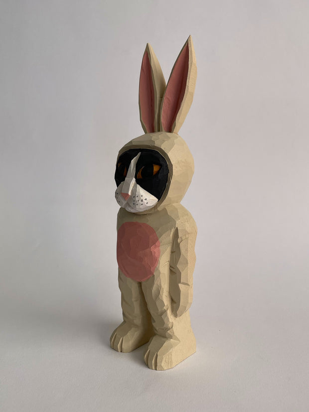 Whittled wooden sculpture of a golden eyed black cat with a white snout wearing a full body light tan bunny costume, with a pink belly and pink ears. It stands up on its legs with its arms straight to its side.