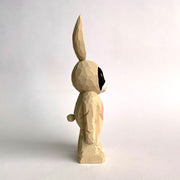 Whittled wooden sculpture of a golden eyed black cat with a white snout wearing a full body light tan bunny costume, with a pink belly and pink ears. It stands up on its legs with its arms straight to its side. It has a small round tail.