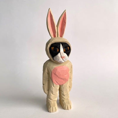 Whittled wooden sculpture of a golden eyed black cat with a white snout wearing a full body light tan bunny costume, with a pink belly and pink ears. It stands up on its legs with its arms straight to its side.