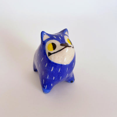 Ceramic sculpture of a blue furred dog, with pointy ears and a very unshapely body. It has yellow eyes and an underbite.