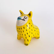 Yellow ceramic sculpture of a dog, with pointy ears and a very unshapely body. It has a blue nose, underbite and black lines drawn on its yellow fur.