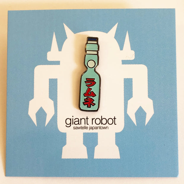 Enamel pin of a Japanese Ramune Soft Drink, a light blue bottle with a white ball towards the top and red Japanese script down the middle. Pin is on a blue backing card.