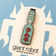 Close up of enamel pin of a Japanese Ramune Soft Drink, a light blue bottle with a white ball towards the top and red Japanese script down the middle.