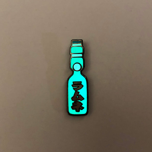Enamel pin of a Japanese Ramune Soft Drink, a light blue bottle with a white ball towards the top and red Japanese script down the middle. Pin is glowing in the dark.