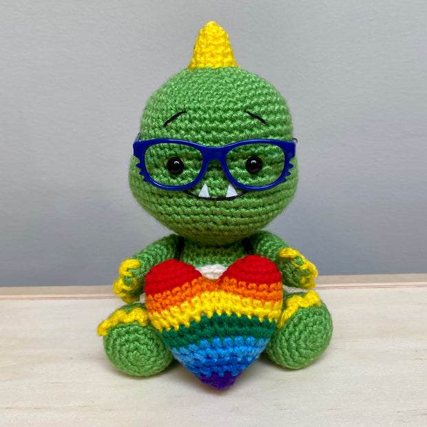 A crocheted sculpture of a chibi Kaiju, green with yellow spikes and nails, wearing blue plastic glasses with a kind expression, smiling with an underbite. It is holding a large rainbow colored heart, also crocheted.  