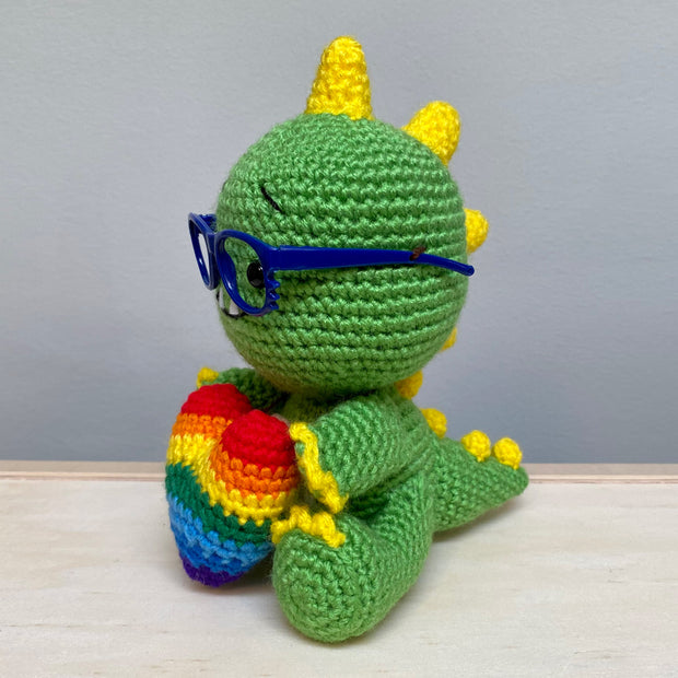 Side view of a crocheted sculpture of a chibi Kaiju, green with yellow spikes and nails, wearing blue plastic glasses with a kind expression, smiling with an underbite.