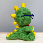 Side view of a crocheted sculpture of a chibi Kaiju, green with yellow spikes and nails, wearing blue plastic glasses with a kind expression, smiling with an underbite.