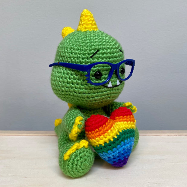 Side view of a crocheted sculpture of a chibi Kaiju, green with yellow spikes and nails, wearing blue plastic glasses with a kind expression, smiling with an underbite.