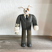 Whittled wooden sculpture of a white dog with a brown spot around his eye, dressed as a business man and standing on two legs like a human.  Its hands are balled into fists and one of them holds a bone.