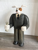 Whittled wooden sculpture of a white dog with a brown spot around his eye, dressed as a business man and standing on two legs like a human. Its hands are balled into fists and one of them holds a bone.