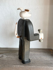 Whittled wooden sculpture of a white dog with a brown spot around his eye, dressed as a business man and standing on two legs like a human. Its hands are balled into fists and one of them holds a bone.