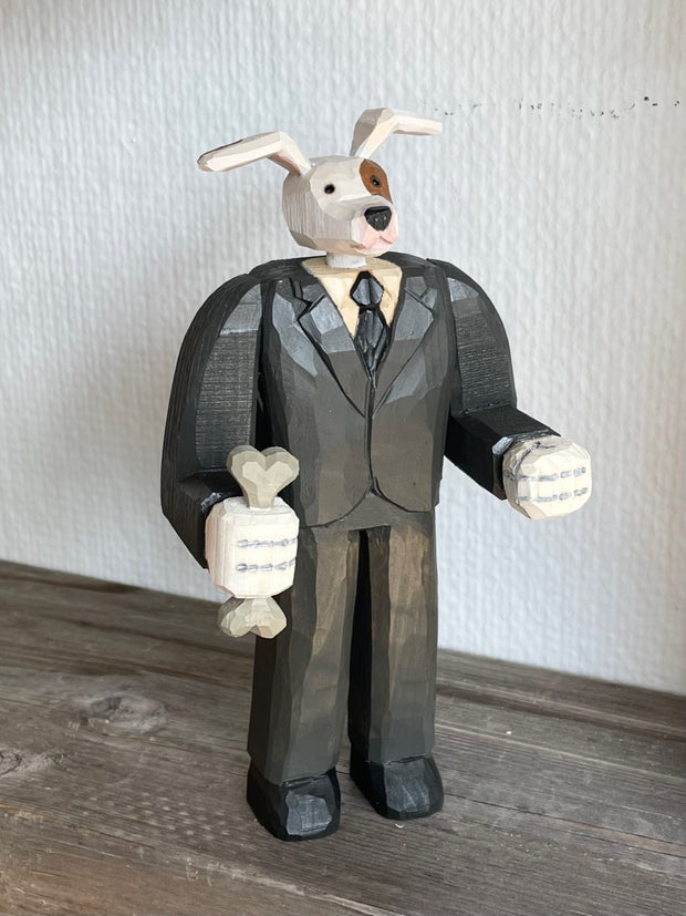 Whittled wooden sculpture of a white dog with a brown spot around his eye, dressed as a business man and standing on two legs like a human. Its hands are balled into fists and one of them holds a bone.