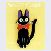 Enamel pin of Jiji with a large red bowtie, sitting and looking head on with an alert expression. On a yellow backing card with Japanese writing.