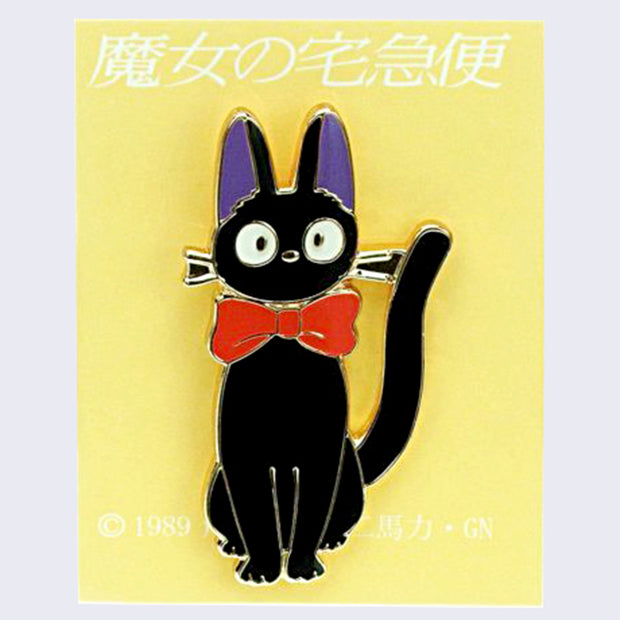 Enamel pin of Jiji with a large red bowtie, sitting and looking head on with an alert expression. On a yellow backing card with Japanese writing.