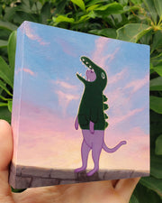 A hand holding a wooden panel painting, posed in front of plants. Painting of an illustrated purple cat with a solemn expression, looking off while wearing a green dinosaur costume that covers their head and upper body. They are standing on a rock wall, with the sunset behind them illuminating their figure.