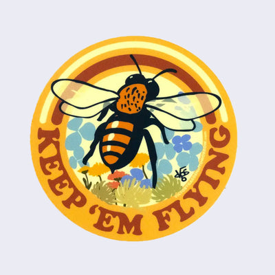 Golden yellow circular sticker with an illustrated honeybee flying over colorful flower background. "Keep 'em flying" is written in stylized bold brown font.