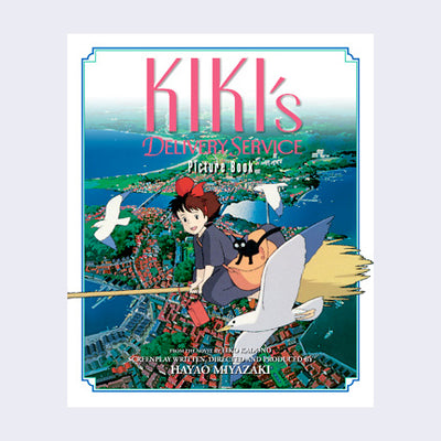 White book cover, featuring an illustration of a girl with a black dress and red bow riding over a sea side town on a broom. A black cat sits on her bag, and they fly among the seagulls.