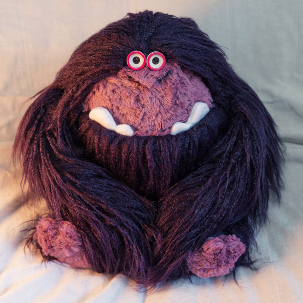 Soft plush sculpture of a sweet, timid looking chubby King Kong, with long dark purple fur and a lighter purple face and feet. It has small button eyes and an underbite showing out of a closed mouth.