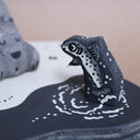 Wooden sculpture of a large whittled sleeping bear, laying on the shore of a lake. A sculpted salmon pops out of the water. Piece is white, black and grey.