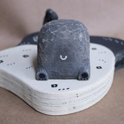 Wooden sculpture of a large whittled sleeping bear, laying on the shore of a lake. A sculpted salmon pops out of the water. Piece is white, black and grey.
