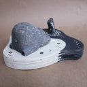 Wooden sculpture of a large whittled sleeping bear, laying on the shore of a lake. A sculpted salmon pops out of the water. Piece is white, black and grey.