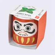 Small red ceramic cup, similar to a sake cup with a cartoon Daruma doll on it. It is encased in a cardboard sleeve that reads "Lucky Plant" over basil leaves and "Sweet Basil Cultivation Kit" written underneath.