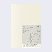 Cream colored blank cover journal within packaging with a gray insert that has Japanese writing and an illustration of a hand holding 3 notebooks.
