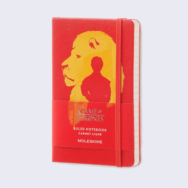Red journal with an elastic closure, cover illustration of a yellow lion bust, looking slightly off to the side. Within the lion is a silhouette of a small figure standing. Paper wrap around insert says "Game of Thrones: Ruled Notebook (Carnet Ligné) Moleskine" 
