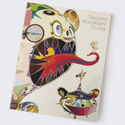 Cover of "Takashi Murakami Prints" book, an abstract Murakami DOB character with many colorful eyes and patterns, sharp dark teeth and a red swirling tongue sticking out. A small character stands on the tongue, with an abstract floating ship like character below.