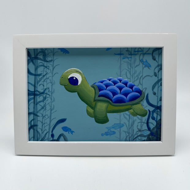Painting of a cartoon style sea turtle, with a blue shell back. It is submerged underwater with other small fish and kelp forests. Piece is framed in thick white wooden frame.