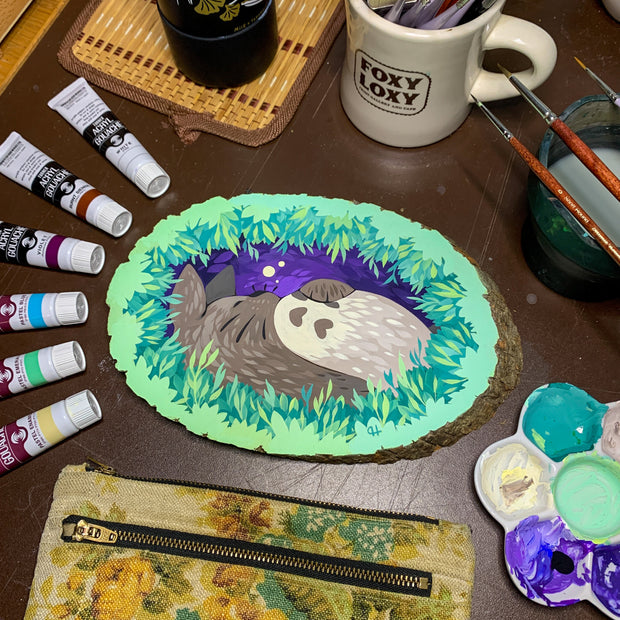 Painting of a sleeping Totoro, framed behind layers of leaves, as if the viewer is looking through a bush to peek on Totoro. Painting is on a wood slice with bark still on the edges. Piece is on a desk with art supplies, showing the creation tools.