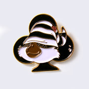 Enamel pin of a fluffy faced creature with a brown snout, wearing a striped sleeping cap that cover its eyes. Character is over a black club.
