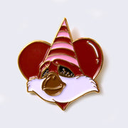 Enamel pin of a fluffy faced creature with a brown snout, and a pink and red striped triangle cap. Character is over a red heart.