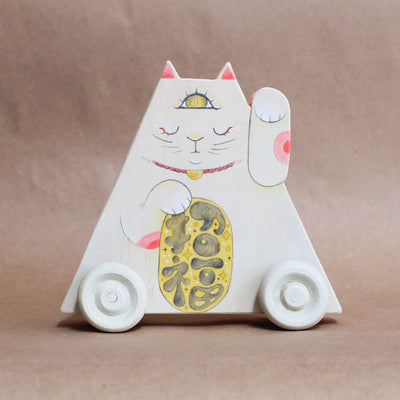 Triangular wooden die cut sculpture of a maneki, a Japanese cat with one paw raised up and another holding a golden token. Its eyes are closed and it has a yellow third eye on its forehead, which is open. Wooden sculpture is on wooden wheels.