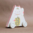 Triangular wooden die cut sculpture of a maneki, a Japanese cat with one paw raised up and another holding a golden token. Its eyes are closed and it has a yellow third eye on its forehead, which is open. Wooden sculpture is on wooden wheels.