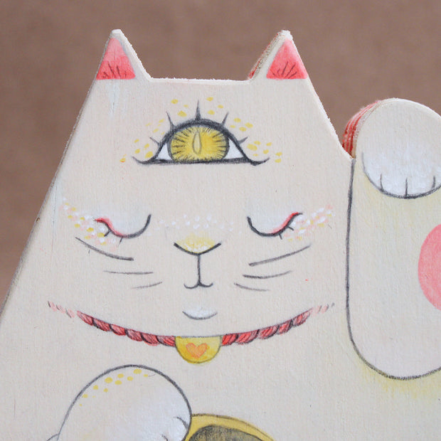Triangular wooden die cut sculpture of a maneki, a Japanese cat with one paw raised up and another holding a golden token. Its eyes are closed and it has a yellow third eye on its forehead, which is open. Wooden sculpture is on wooden wheels.