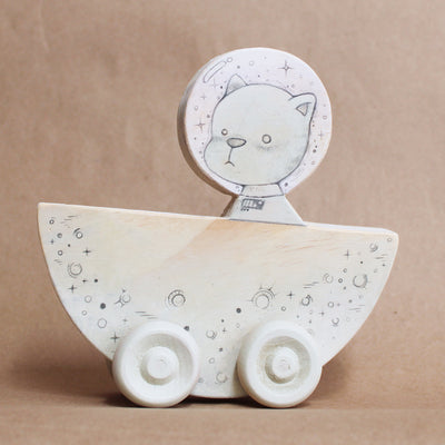 Wooden die cut sculpture of a cat wearing a space helmet, riding in a half moon with wheels on it. Details of the piece are drawn in with pencil.