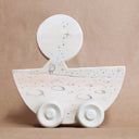 Wooden die cut sculpture of a cat wearing a space helmet, riding in a half moon with wheels on it. Details of the piece are drawn in with pencil.