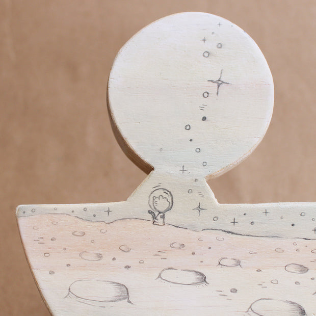Wooden die cut sculpture of a cat wearing a space helmet, riding in a half moon with wheels on it. Details of the piece are drawn in with pencil.