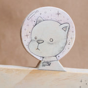Wooden die cut sculpture of a cat wearing a space helmet, riding in a half moon with wheels on it. Details of the piece are drawn in with pencil.