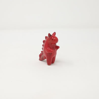 A small ceramic red Godzilla figure with two devil horns on its head. It has minimal facial and body details. It has pointed spikes on its back and is standing with its short arms extended.