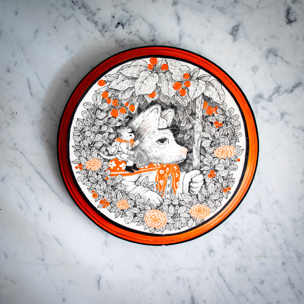 Black ink illustration on white paper with orange color accents of a dog, appearing from a circular framing of leaves and flowers. It holds up a stick like a staff, and has a small cat in an explorer's outfit riding atop its shoulder. On a orange circle panel.