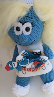 Close up view of a crocheted plush sculpture of a smiling Smurfette, holding a bowl filled with various sewn Smurfs that appear flat like gingerbread cookies.