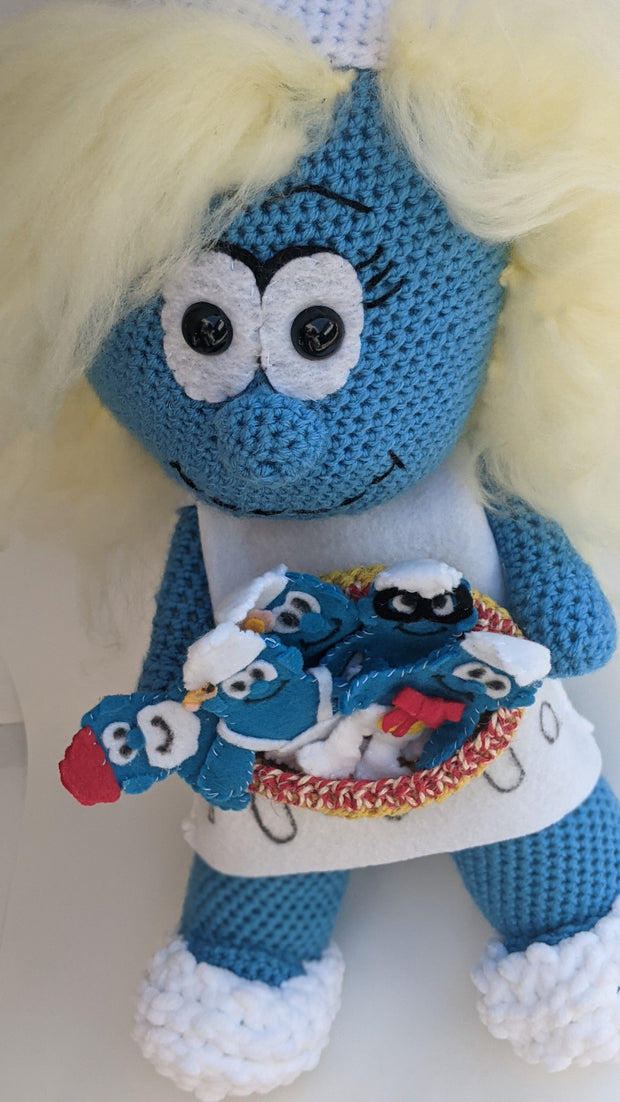 Close up view of a crocheted plush sculpture of a smiling Smurfette, holding a bowl filled with various sewn Smurfs that appear flat like gingerbread cookies.