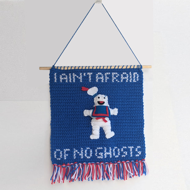 A crocheted banner attached to a skinny wooden stick, hanging on a wall. Banner is blue with red, white and blue threads at the bottom. A sewn Stay Puft Marshmallow Man is in the middle of the banner between writing that reads "I Ain't Afraid of No Ghosts" crocheted in all caps.